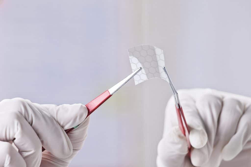 gloved hands holding square of graphene with tweezers